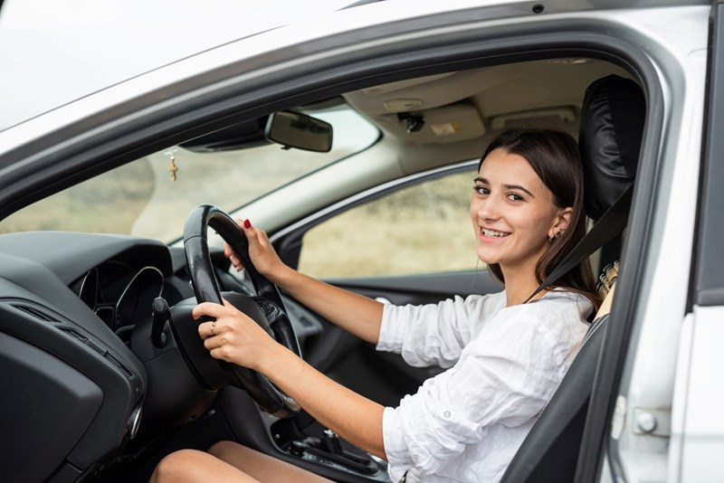Smiling woman driving car, looking at camera with hands on steering wheel and seatbelt on.