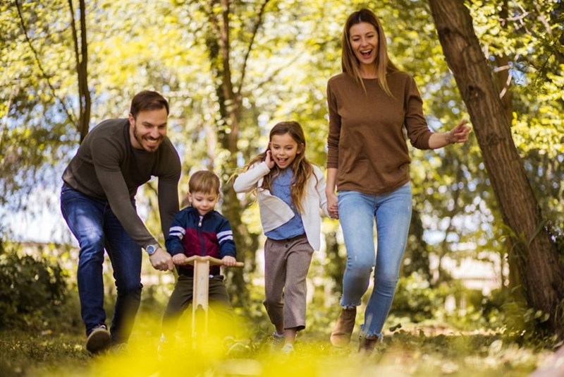 Parents and 2 kids play outdoors with trees in the background.