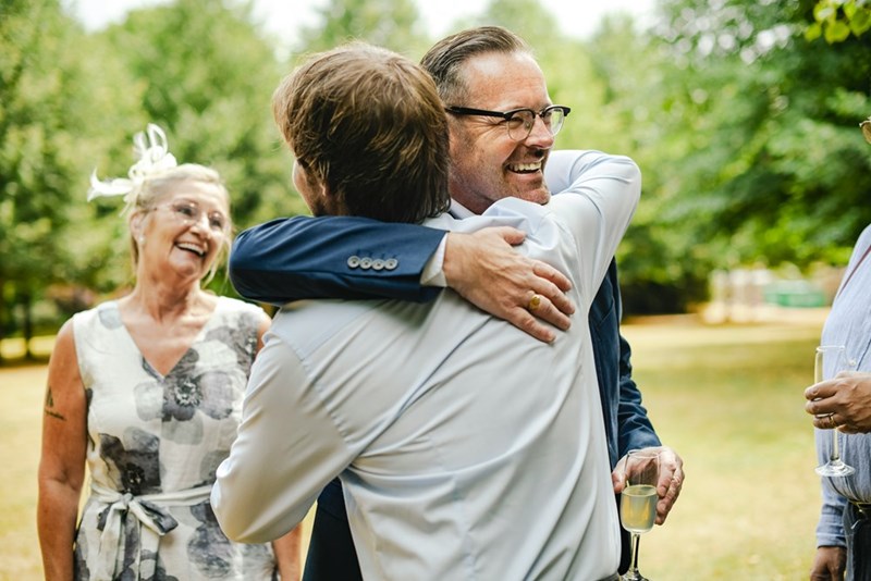 An older male wedding guest hugs another guest during an outdoor ceremony.