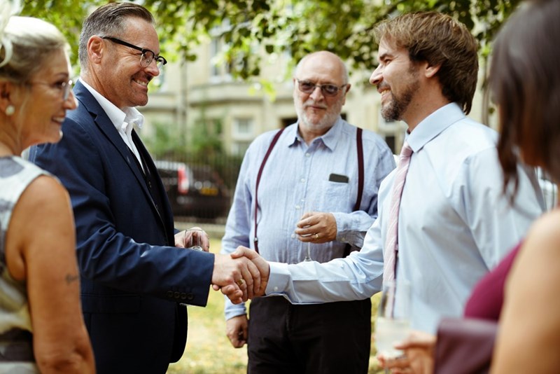 An older male wedding guest shakes hands with another guest in a social circle during an outdoor reception.