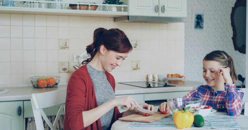 A mom sits at a kitchen table, cutting tomatoes while her daughter watches