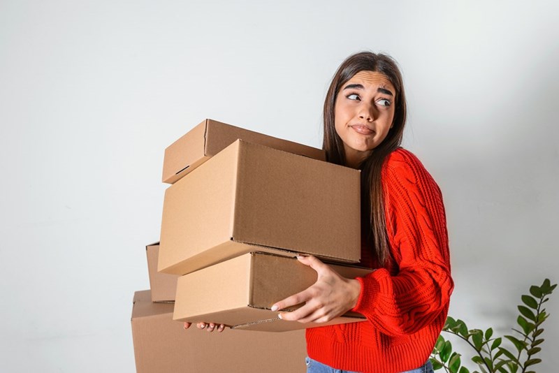 A woman looks around at an empty space as she carries packed cardboard boxes.