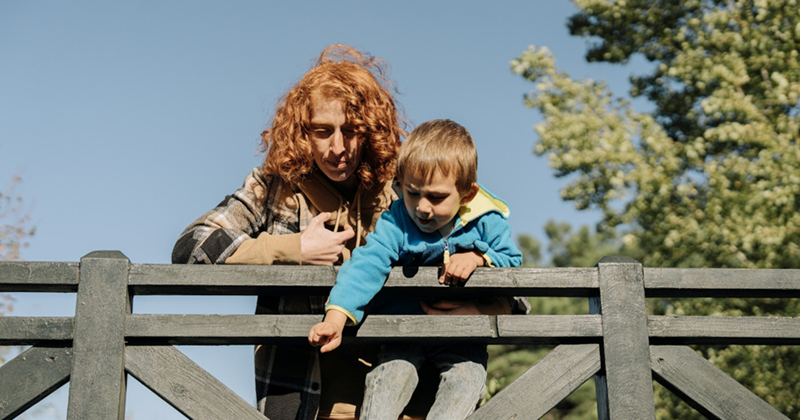 A woman and a young boy lean over a wooden railing on a sunny day