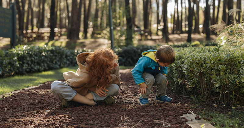 A woman sits cross-legged beside a young boy, crouching down to look at something in a forest