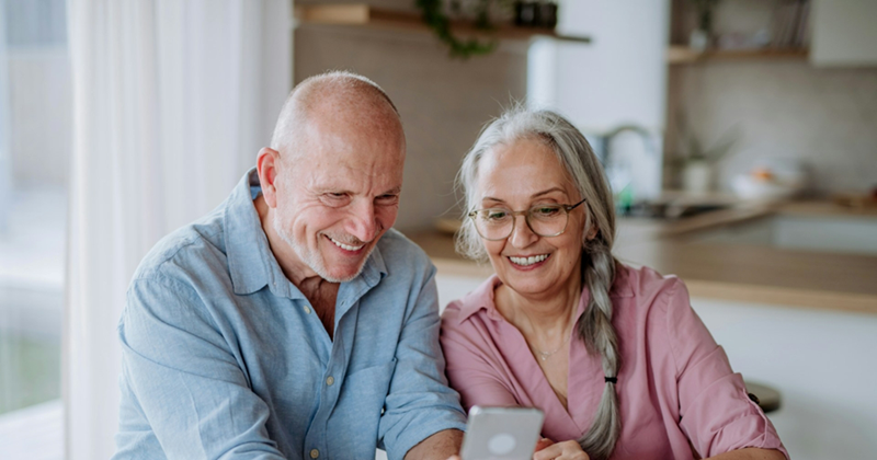 An older couple smiles while sitting together and looking at a cellphone