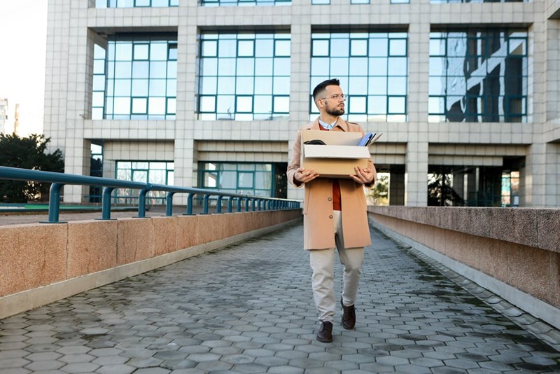A wider shot of a businessman holding a cardboard box of personal items as he walks outside of an office building.