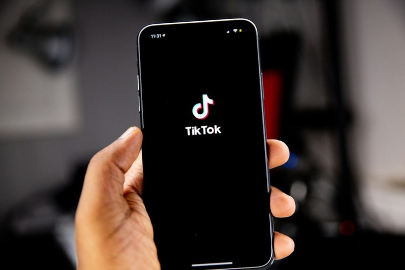 A hand holds a smartphone displaying the TikTok logo against a dark blurred background, highlighting the social media platform at the center of the controversy.