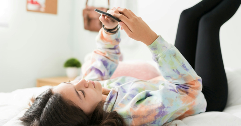 A teenage girl lies on her bed with her legs on the wall, looking at her cellphone