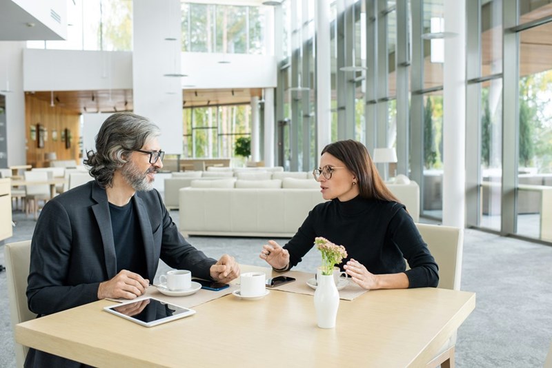 Two confident colleagues in smart casualwear and eyeglasses discussing financial news or projects of their company by cup of coffee