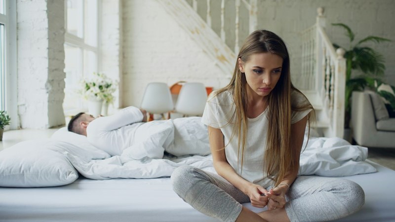 A woman sits on the edge of the bed upset with a man curled up behind her