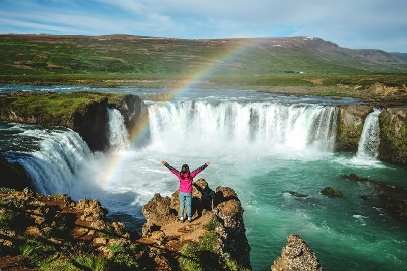 Tourist stands at the top of a waterfall in Iceland with a rainbow appearing above her.