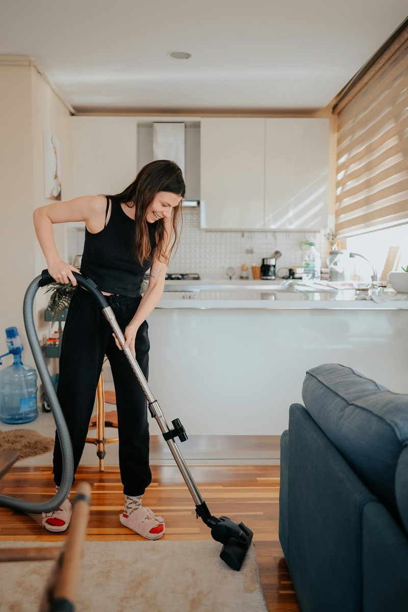 Woman vacuuming her apartment wearing black joggings and slippers.