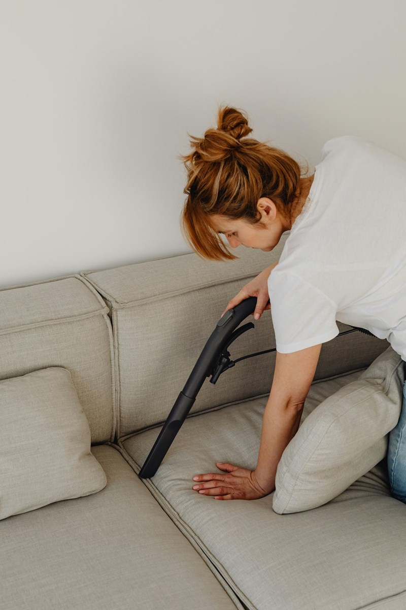 Woman wearing a white shirt and a bun vacuuming her beige couch.
