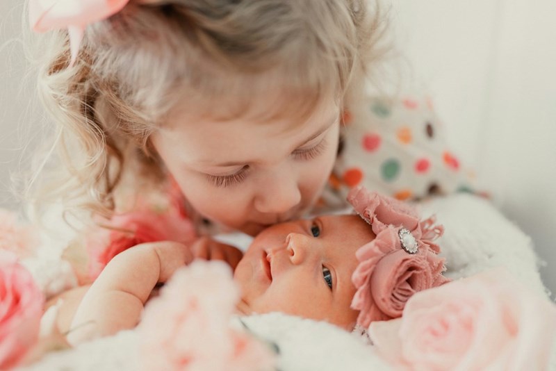 Older sister kisses newborn baby sister surrounded by flowers