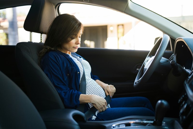 A pregnant woman adjusts her seatbelt in the driver's seat of her car.