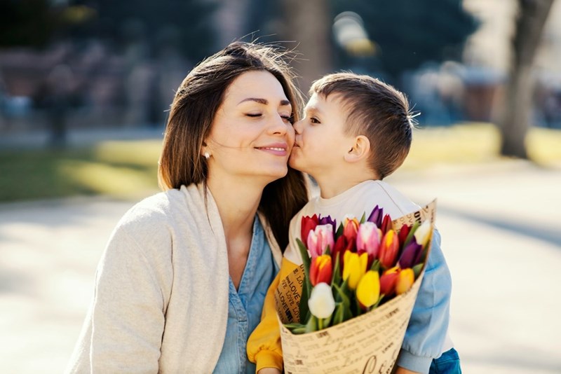 A young child sweetly kisses his smiling mother on the cheek while holding a colorful bouquet of flowers outdoors for Mother’s Day.