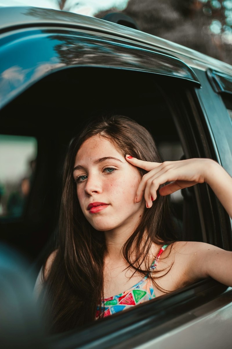 A girl rests her arm on an open car window, touching her face with her hand