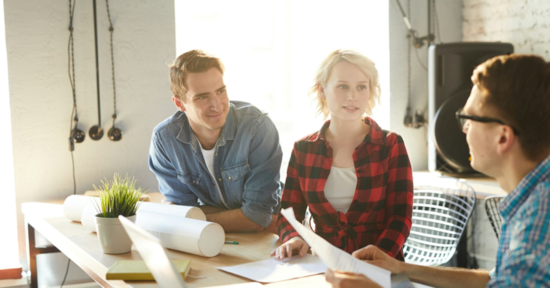A male and female coworker sit beside one another at a table, listening to a coworker holding a piece of paper speak