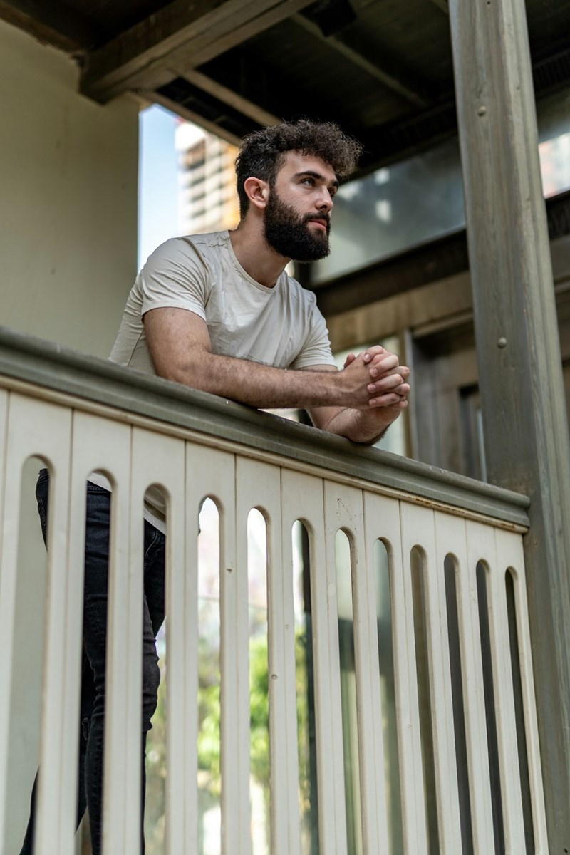 Bearded man standing on a balcony with clasped hands, gazing thoughtfully outside a modern building.