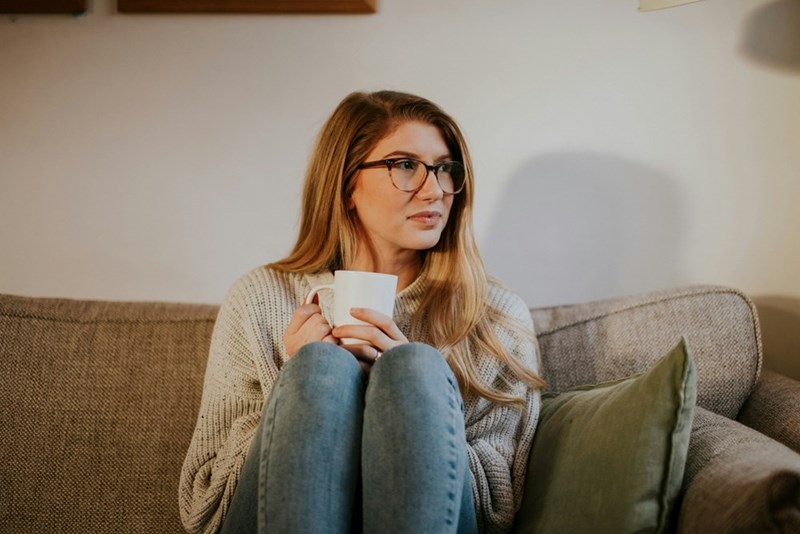 Woman wearing glasses relaxing on a couch with a coffee mug, cozy sweater, and warm indoor lighting.