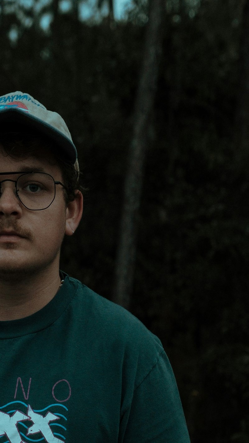 Young man in glasses and graphic sweatshirt posing outdoors against a dark wooded background.