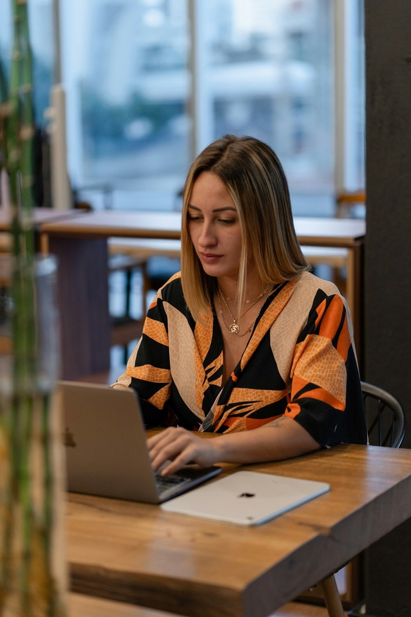 Woman typing on a laptop at a wooden table in a stylish café workspace with soft natural lighting.