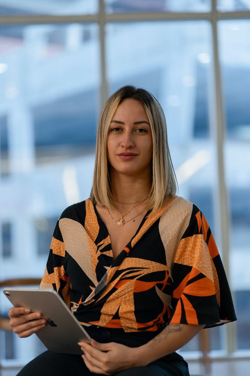 Woman holding a tablet indoors with large windows behind her, wearing a patterned blouse and layered necklaces.