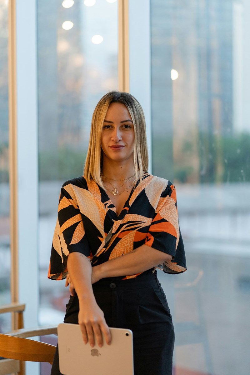Woman holding a tablet indoors near tall windows, wearing a patterned blouse and layered gold necklaces.