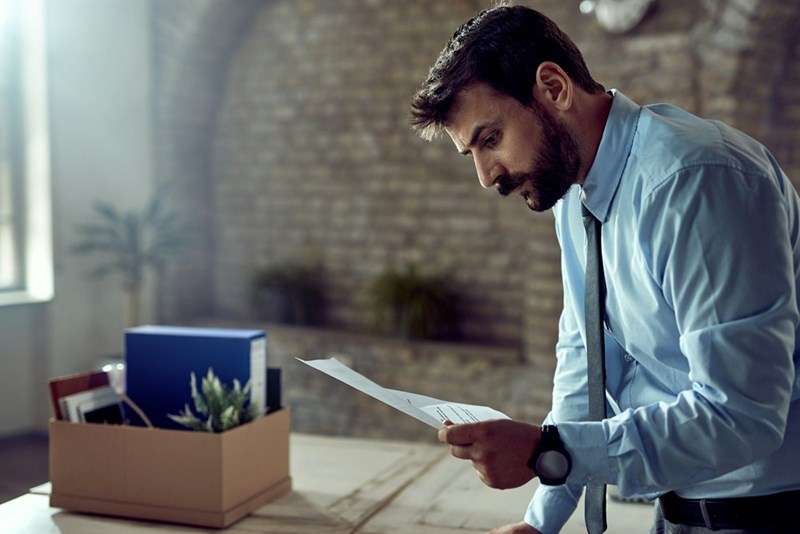 A stressed office employee stands at his desk reading paperwork beside a cardboard box, seemingly reacting to bad news at work.