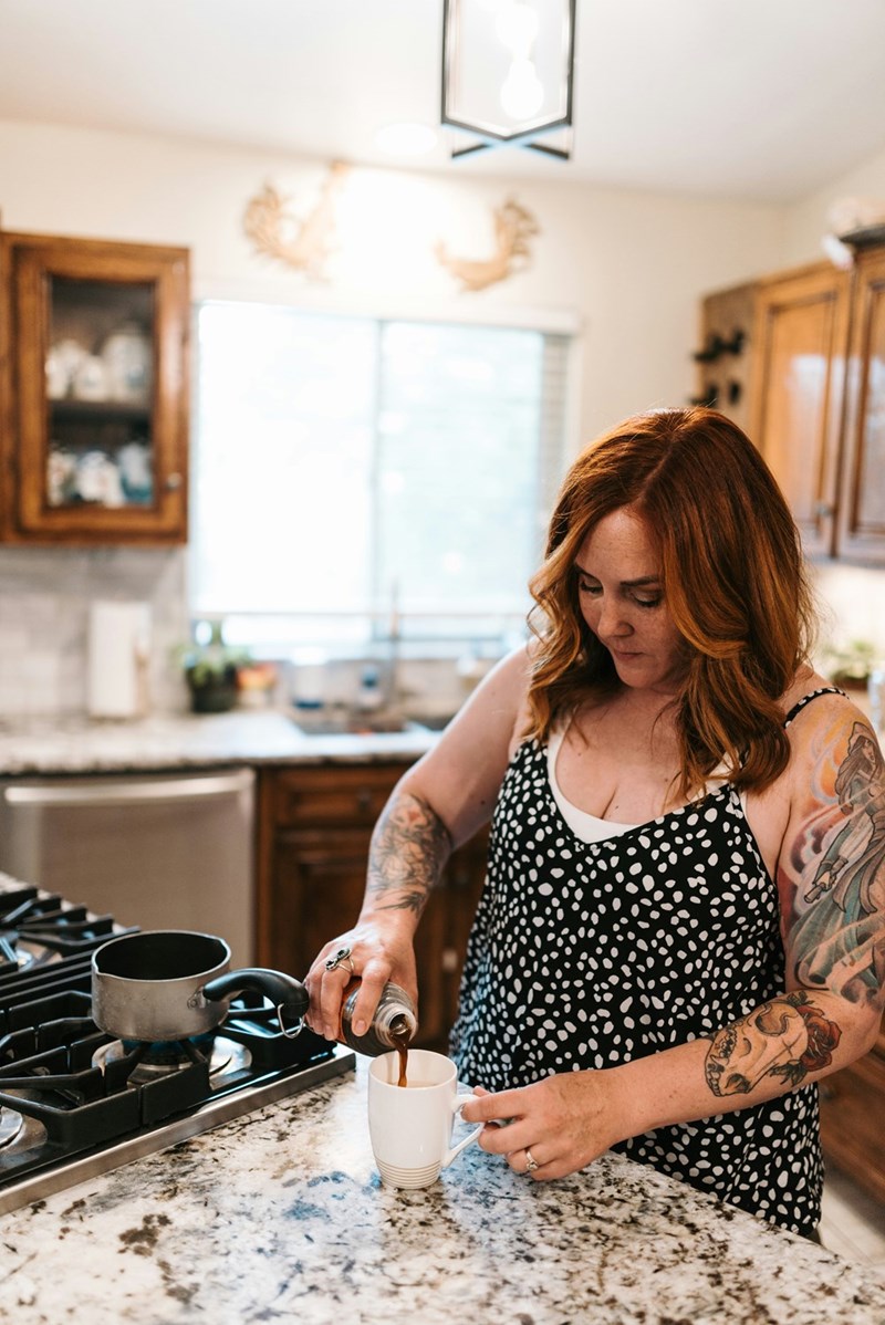 Woman with tattoos pouring coffee into a mug at a kitchen counter beside a stovetop.