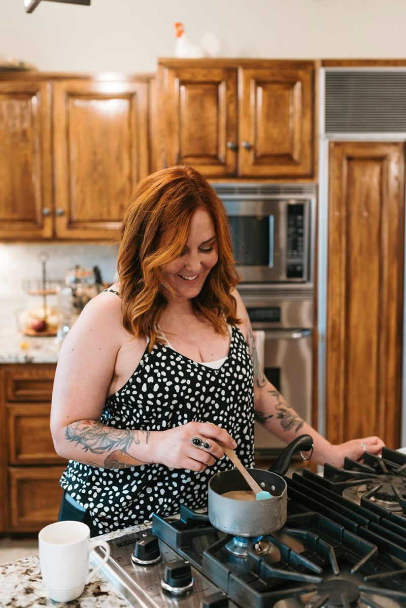 Woman with tattoos stirring a saucepan on a gas stove in a warm wood-paneled kitchen.