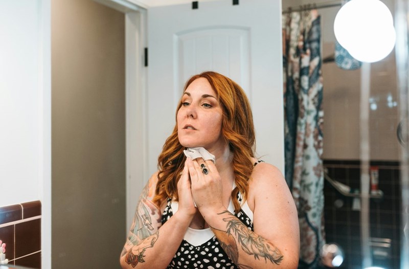 Woman with tattoos applying skincare to her neck while standing in a bathroom mirror.