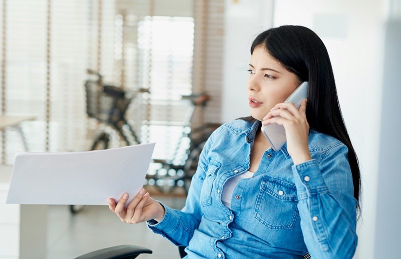 Woman talking on a smartphone while reviewing paperwork in a bright home.