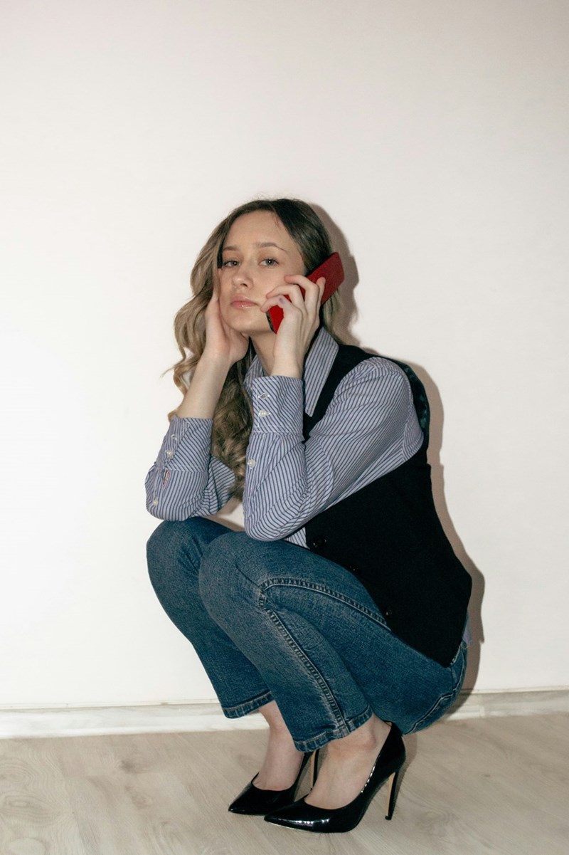 Woman in jeans and heels crouching while talking on a smartphone indoors against a plain wall.