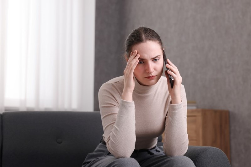 Worried woman sitting on a couch holding a smartphone with her hand on her forehead.