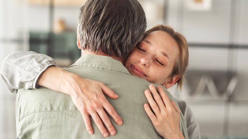 Woman hugging an older man with eyes closed in a close emotional embrace indoors.