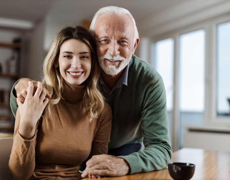 Young woman and older man smiling together indoors with their arms around each other.