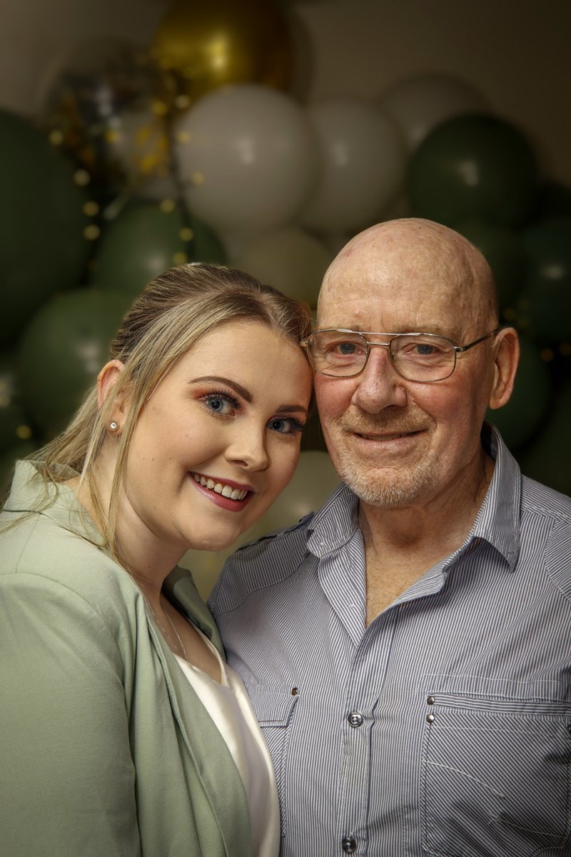 Young woman and older man smiling together indoors with decorative balloons in the background.