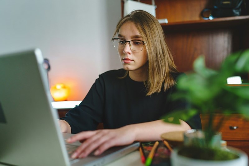 Woman in glasses working on a laptop at a desk with a plant and shelves in the background.