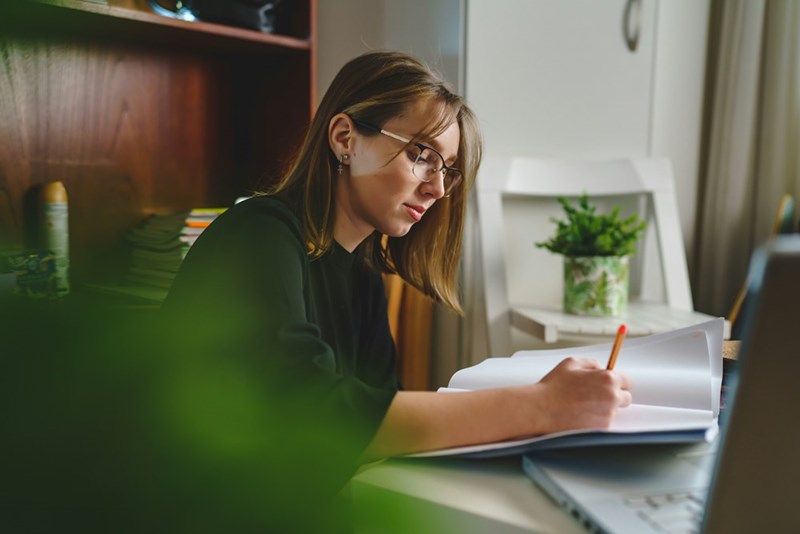 Woman in glasses writing in a notebook at a desk beside a laptop.