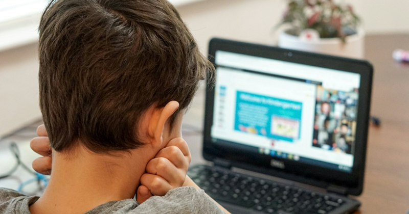 The back of a boy's head as he sits at a table and looks at a blurred laptop screen