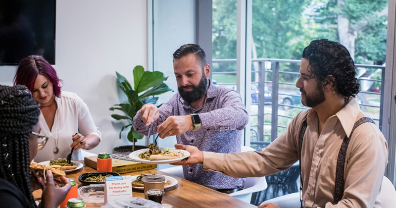 A man holds up a plate of food for his coworker to take from as their other coworkers eat