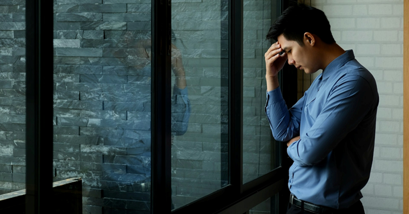 A man holds his head in his hand as he stands by a large window looking out on a wall