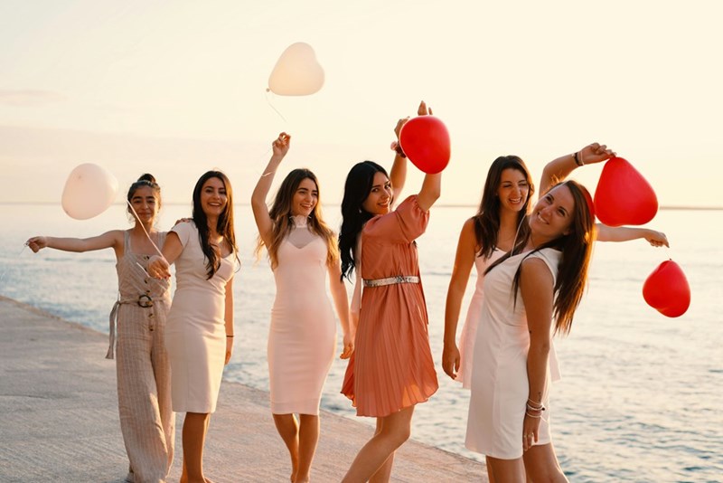 6 women in a bachelorette party hold heart-shaped balloons while enjoying a beach walk.