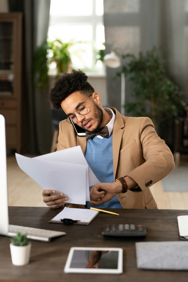 Stressed employee checks his watch while also being on the phone and checking paperwork.Stressed employee checks his watch while also being on the phone and checking paperwork.