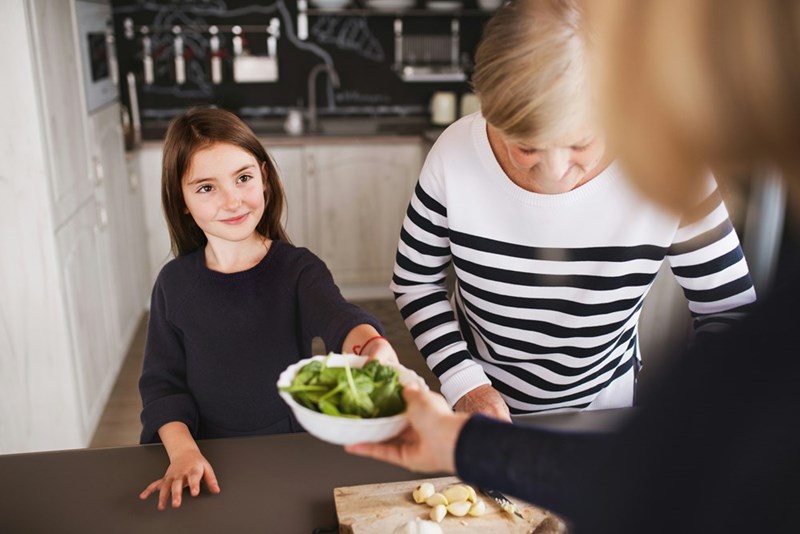 Young girl is handed a bowl of spinach leaves while preparing a meal.