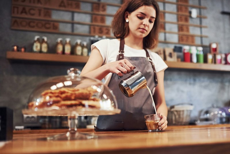 Woman wearing an apron working behind a bar serving coffee.