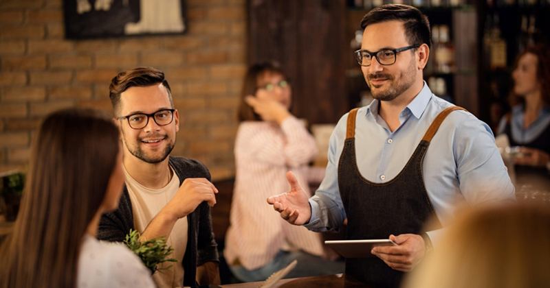 A male server holding a tablet speaks to a couple sitting at a table