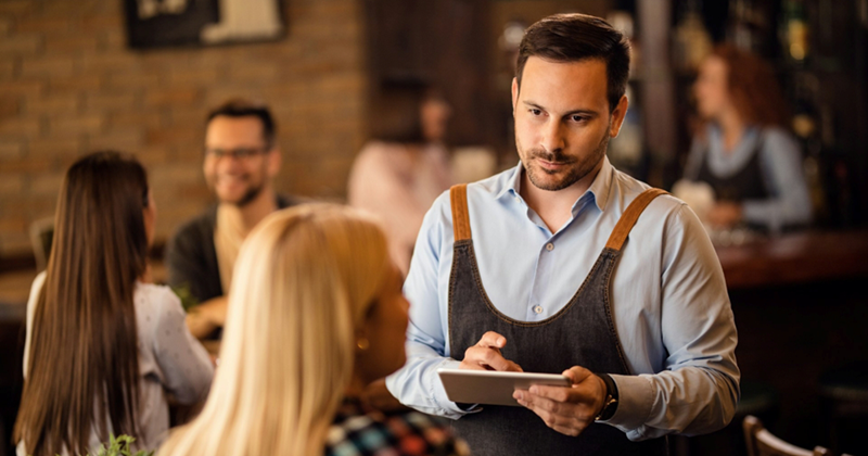 A male server holding a tablet takes a table's order
