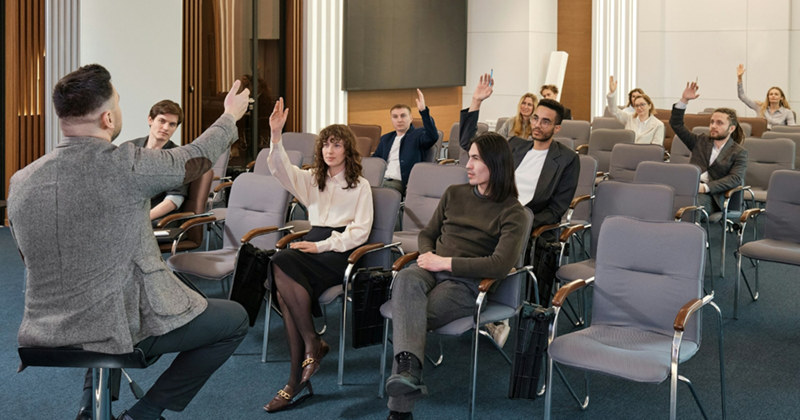 A college teacher sits on a stool and takes questions from students seated in front of him